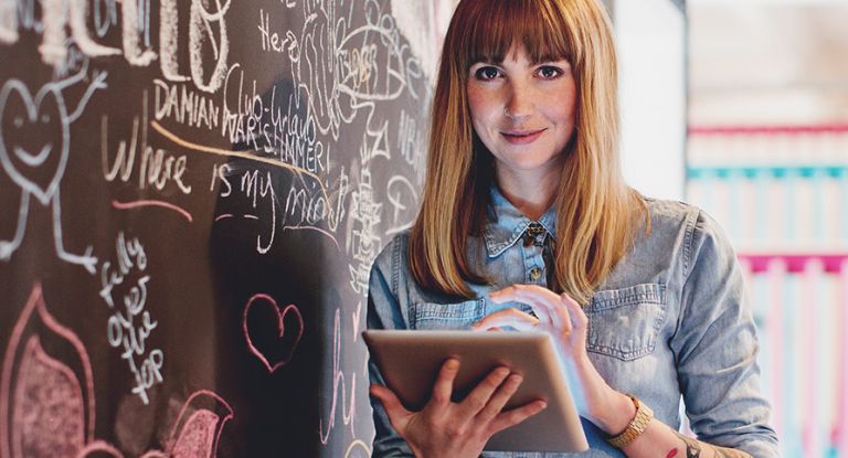 Frau mit Tablet Junge Frau steht mit Tablet in der Hand neben einer bunt bemalten Kreidewand und lächelt dabei freundlich in die Kamera.