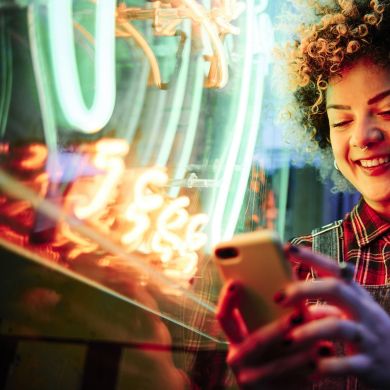 Frau mit Handy vor Neonröhre Frau mit lockigen kurzen Haaren steht vor einem Neonlicht und schaut lächelnd auf ihr Handy