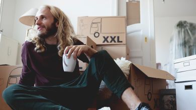 Junger Mann mit weißer Tasse in der linken Hand. Sitzt auf dem Boden in einer Wohnung vor ein paar gestapelten Umzugskartons.