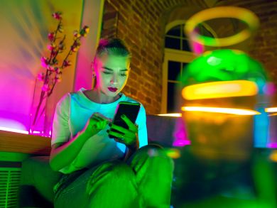 Frau sitzt mit Handy auf der Couch Eine Frau sitzt mit einem Handy auf der Couch und ist umgeben von Neonlichtern