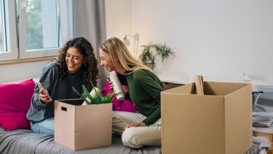 Zwei Frauen ziehen um Zwei Frauen sitzen lächelnd auf einem Bett mit Umzugskartons