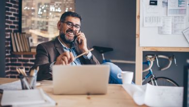Mann vor Laptop Mann mit Brille sitzt lachend vor einem Laptop am Schreibtisch, hält dabei einen Stift in der linken und ein Handy am Ohr in der rechten Hand.