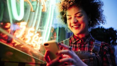 Frau mit Handy vor Neonröhre Frau mit lockigen kurzen Haaren steht vor einem Neonlicht und schaut lächelnd auf ihr Handy